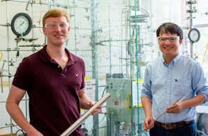 Kevin Barnett and Kefeng Huang stand in front of a reactor that is encased in glass