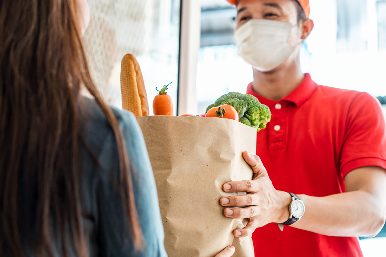 Asian delivery man wearing face mask in red uniform handling bag of food, fruit, vegetable give to female costumer in front of the house